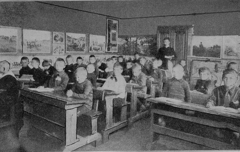 A teacher standing at the back of a full classroom of pupils at wooden desks, from a 1916 book